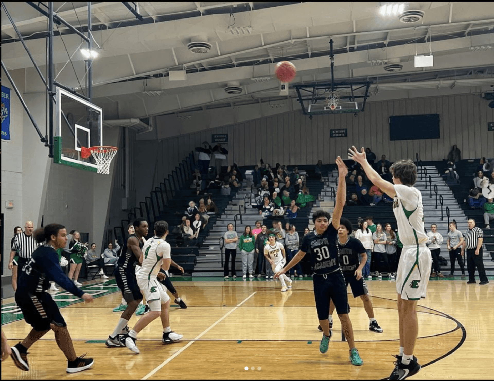 High school basketball game, player in white uniform shooting a three-pointer surrounded by opponents.