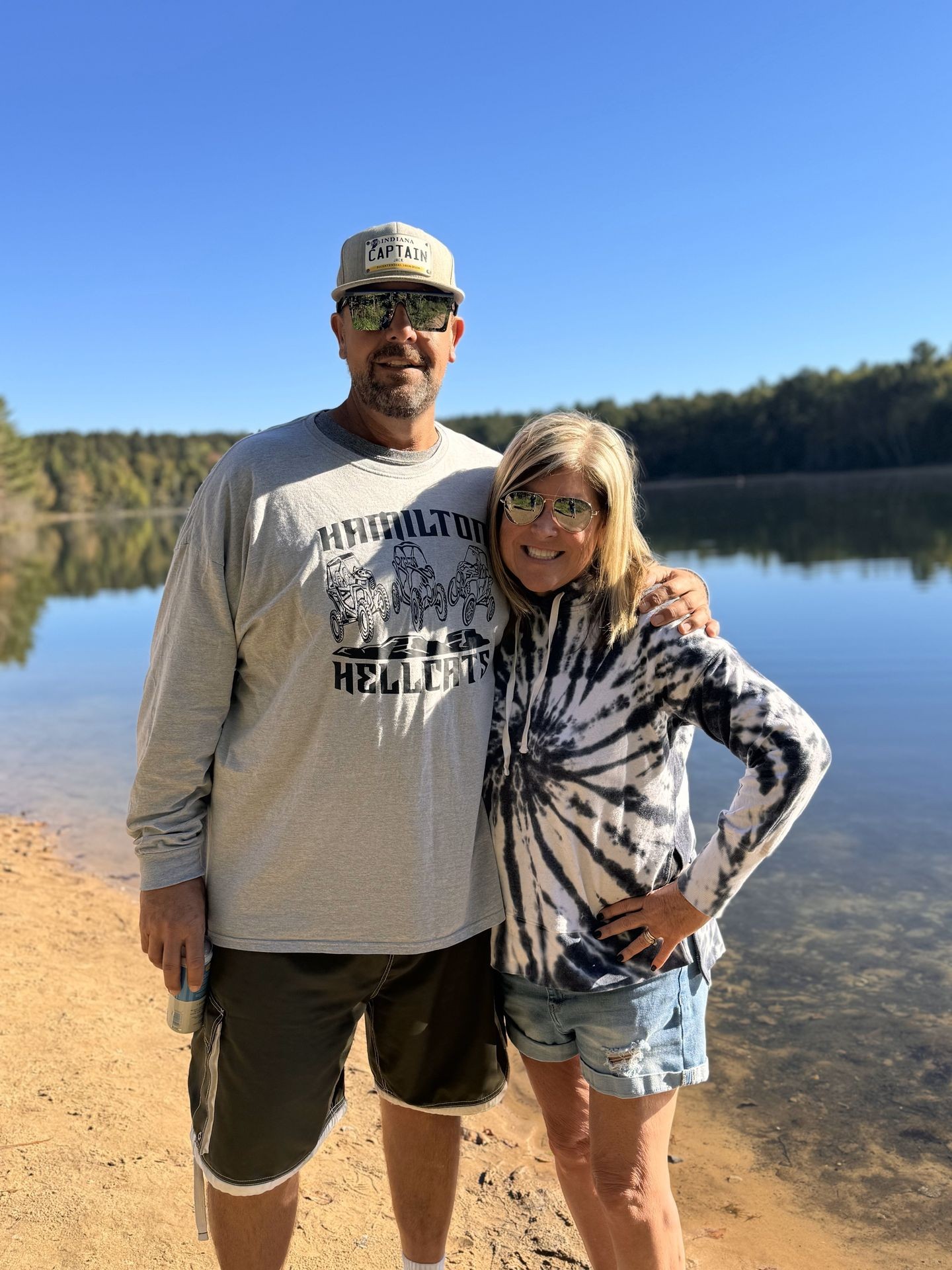 Two people standing close together by a lake, with trees and clear blue sky in the background.