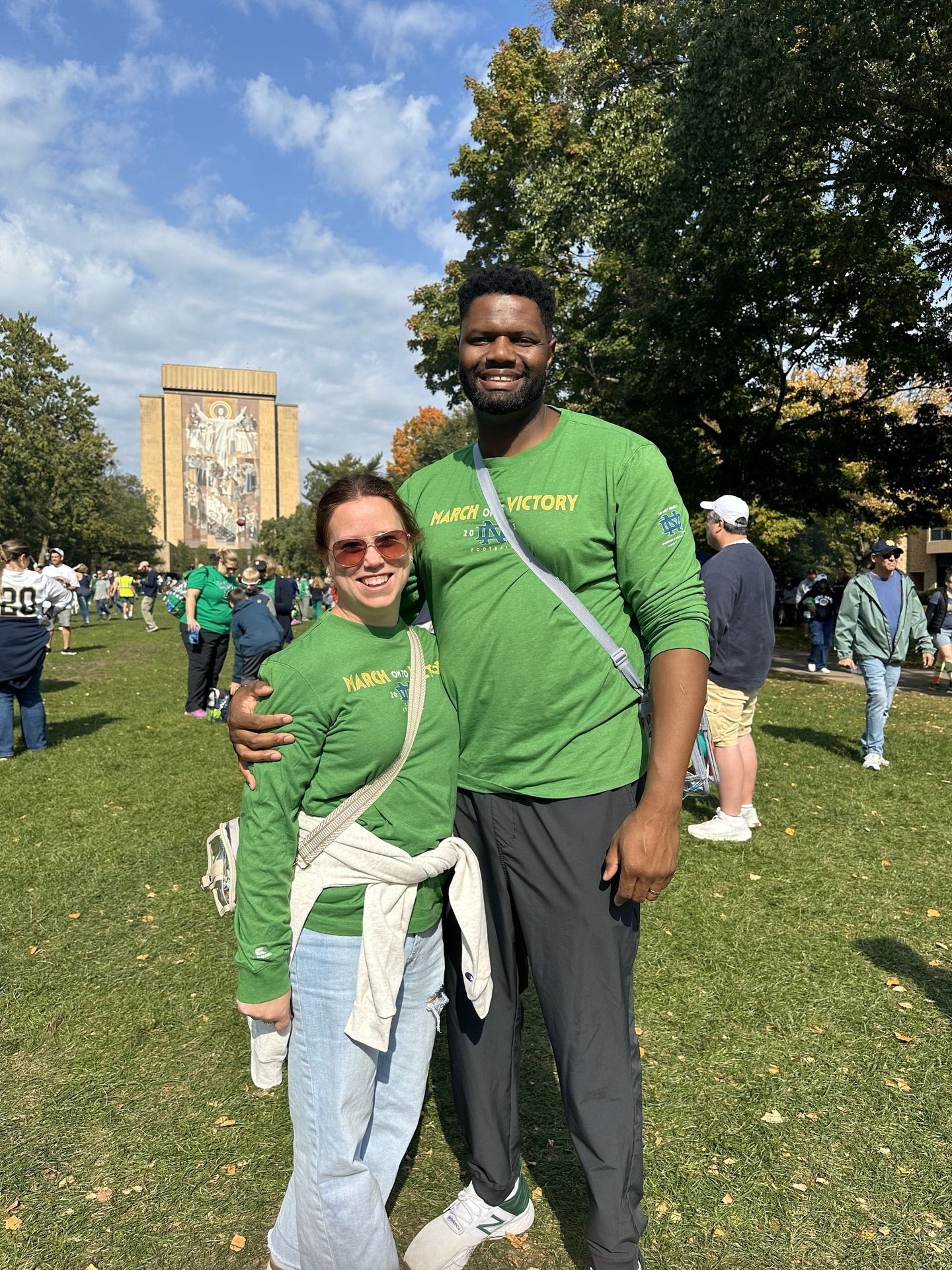 Two people in green shirts smiling on a grassy field with a mural in the background.