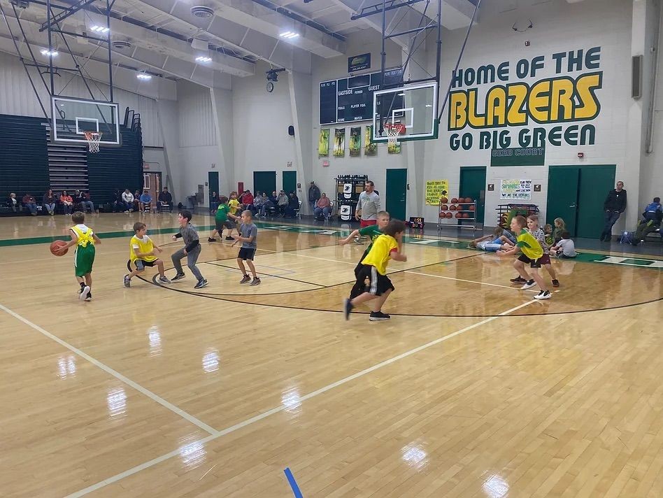 Children playing basketball in a gym with spectators watching from bleachers.