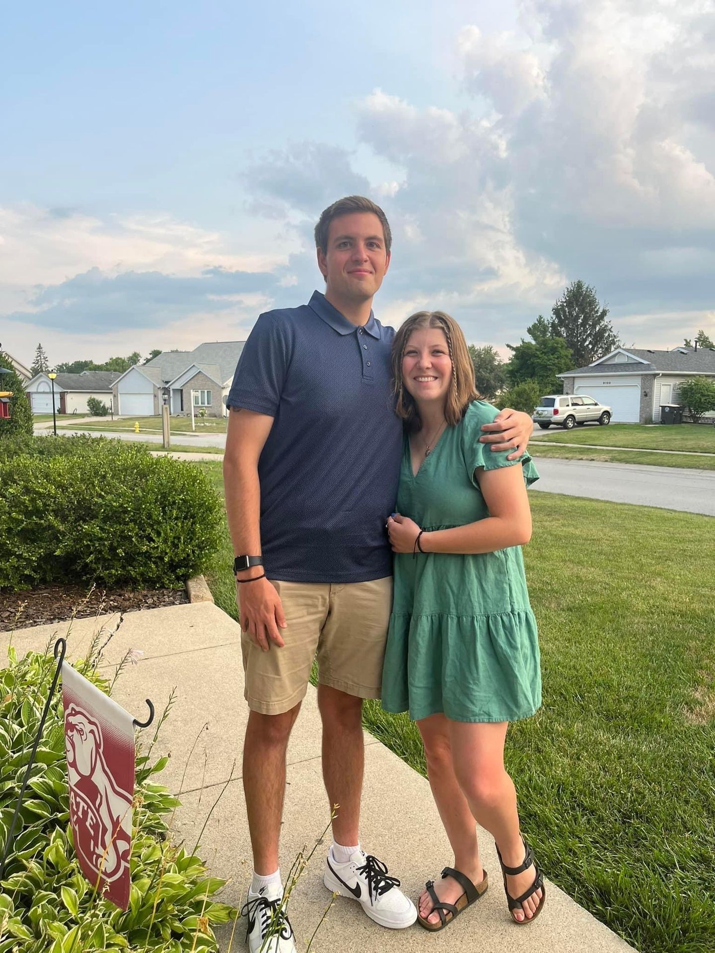 Smiling couple standing outside on a sidewalk with houses and a cloudy sky in the background.