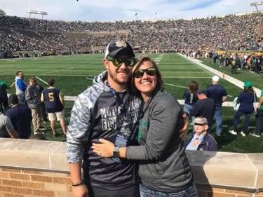 Couple embracing and posing by a wall in a stadium filled with spectators watching a football game.